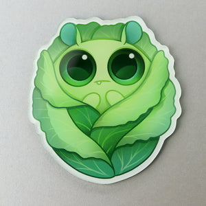 A cute, cartoonish green caterpillar nestled within the folds of a lettuce leaf, designed as a playful sticker with big, expressive eyes and a gentle smile by Amanda Spayd on a "Cabbage" Acrylic Magnet from Bindlewood Shop.