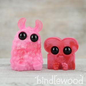 Two whimsical, cartoon-style resin figures with big black eyes, one pink and rabbit-like known as Strawberry Sparkle Crème PomPom and the other red, resembling toast or a character with ears from the Valentines Day Set by Bindlewood Shop.