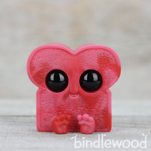 A whimsical, pink, Cherry Cordial Toastboy-shaped character with large, expressive black eyes and tiny feet holding a heart.