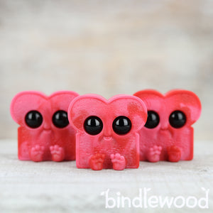 Three adorable Cherry Cordial Toastboy pink toy figures with large black eyes, resembling cartoonish hearts, lined up against a soft-textured background by Chris Ryniak.