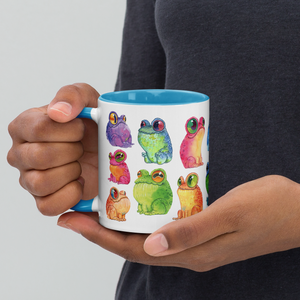 A person holding a Frog Frenzy Mug adorned with colorful illustrations of whimsical frogs inspired by Chris Ryniak watercolor frog paintings from Bindlewood Shop.