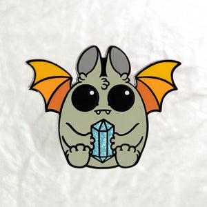 Illustration of a cute cartoon bat from Thimblestump Hollow with large eyes and orange wings, holding an Atticus Enamel Pin from Bindlewood Shop. The background is a crumpled white texture.