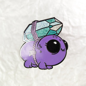 A Gemscuffler Enamel Pin featuring a cute, cartoon-style purple dragon with large, shiny turquoise and blue scales, a big black eye, and delicate white details from Bindlewood Shop, displayed against a soft
