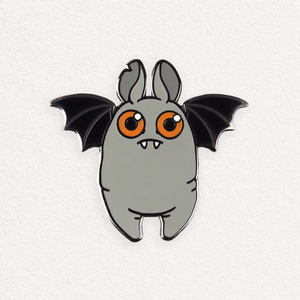 A hard enamel pin of a cartoon-style, gray fat bat character with large orange eyes, small fangs, and black wings, set against a white background. (Bat (Orange Eyes) Pin by Bindlewood Shop)