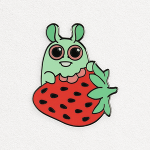 Illustration of a cute, green cartoon character with large eyes and an antenna, wearing a red Strawberry Bunny Pin costume dotted with seeds. The character joyfully holds a green leaf, designed by Amanda Louise Spay for Bindlewood Shop.