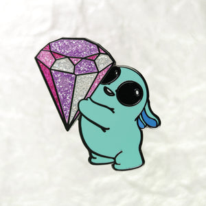 A cute cartoon-style illustration of a teal bunny holding a large, sparkling, purple and silver Topaz Enamel Pin from Bindlewood Shop, all set against a crinkled white background.