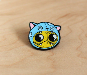 A colorful enamel Catstume Pin featuring a whimsical design of a cat costume with big, yellow eyes, blue fur, and pink ears, adorned with stitches and gears, suggesting a mechanical or steampunk style from Bindlewood Shop.