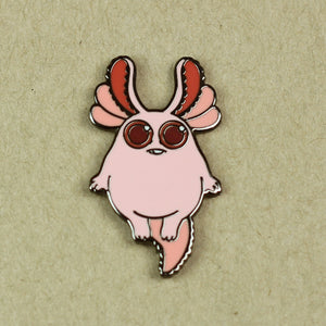 A whimsical Tiny Axolotl-bunny Pin featuring a pink creature with large red eyes and curved antlers against a beige background by Bindlewood Shop.