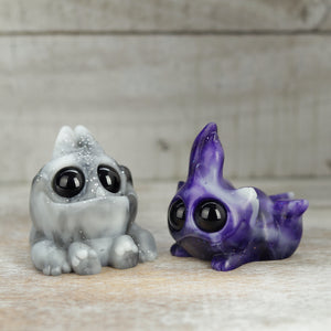 The Thundersparkle Tank & Mooncloud Bugbite Set from Bindlewood Shop features two glow-in-the-dark resin figures—a marbled gray frog and a purple, long-eared rabbit with glossy black eyes—displayed on a wooden surface.