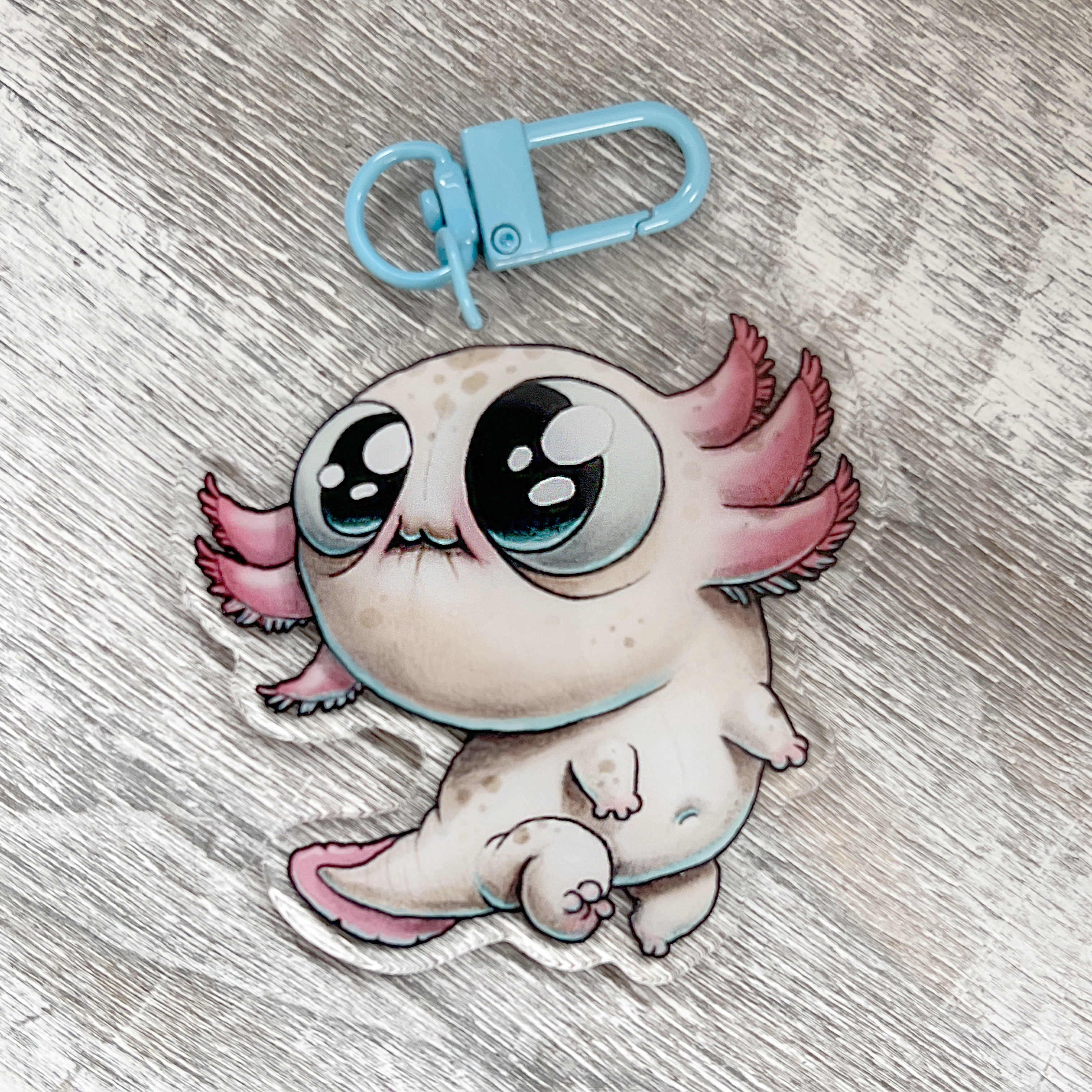 A charming Axolotl Keychain from Bindlewood Shop, featuring big, shiny eyes, pink gills, and a small light blue clasp, rests on a textured grey surface. Crafted in the whimsical style of Chris Ryniak, this delightful acrylic keychain makes a perfect Axolotl charm for your collection.