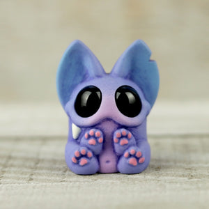 A small, purple, bat-like figurine with large black eyes and a single visible fang, created by Chris Ryniak, sits upright with its tiny paws held close to its face. The background is a blurred, light-colored surface reminiscent of the vibrant atmosphere at Five Points Festival.