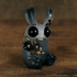 The Amanda L Spayd Black & Gold Peppercorn resin figurine is a small, dark gray, rabbit-like creature featuring large black eyes, tiny fangs, and hand-painted gold-accented black flowers adorning its body. Set on a wooden surface against a softly blurred brown background, this charming piece embodies an enchanting and whimsical allure characteristic of Amanda L Spayd’s unique style.