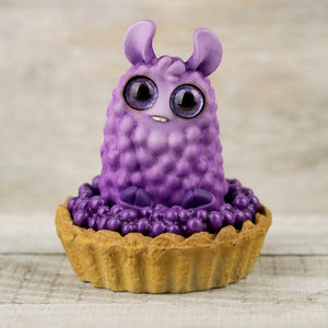 A small, cute figurine of a purple, fluffy creature with big round eyes, tiny ears, and little feet sits inside a tart shell filled with purple berries. This special edition piece is hand-painted and perfect for fans of unique collectibles. Introducing the Blueberry Tart Pom Pom from Bindlewood Shop! The background is a wooden surface.