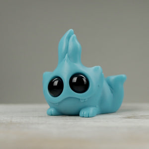 A small blue opaque resin figure of a cute creature with large black eyes, chubby cheeks, and a playful expression. Created by Chris Ryniak, the Robin's Egg Blue Bugbite has a fin-like structure on top of its head and appears to be lying down on a flat surface. The background is plain and neutral.