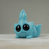 A small blue opaque resin figure of a cute creature with large black eyes, chubby cheeks, and a playful expression. Created by Chris Ryniak, the Robin's Egg Blue Bugbite has a fin-like structure on top of its head and appears to be lying down on a flat surface. The background is plain and neutral.
