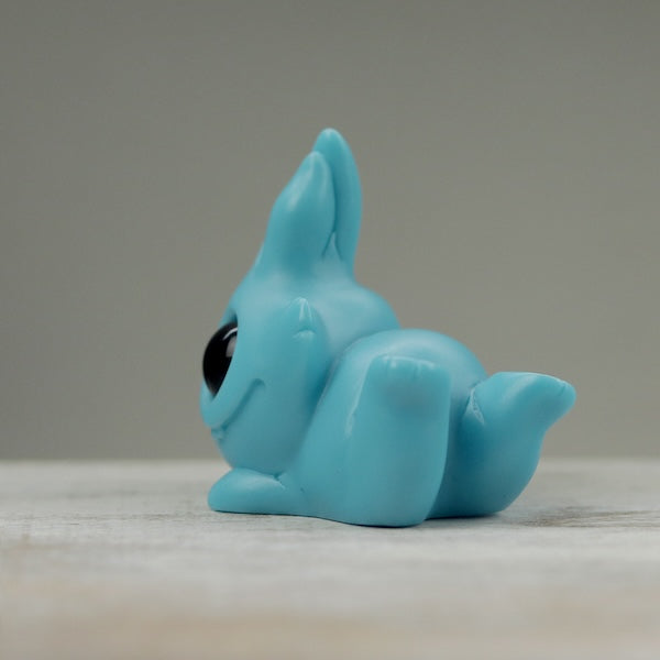 A Robin's Egg Blue Bugbite by Chris Ryniak, a small blue opaque resin figure of a creature with pointed ears and large black eyes, is lying on its stomach facing to the side. Created by Chris Ryniak, the figurine has a rounded body with legs tucked underneath and a tail sticking up. The background is plain and gray.