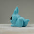 A Robin's Egg Blue Bugbite by Chris Ryniak, a small blue opaque resin figure of a creature with pointed ears and large black eyes, is lying on its stomach facing to the side. Created by Chris Ryniak, the figurine has a rounded body with legs tucked underneath and a tail sticking up. The background is plain and gray.