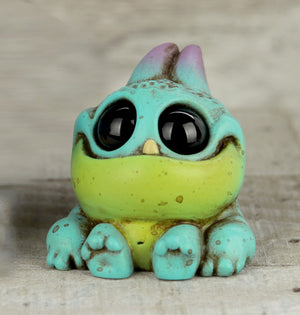 Peacock Tank by Chris Ryniak is a small, chonky resin figure of a colorful cartoonish frog with big black eyes, a wide yellow-green mouth, teal body, tiny feet, and purple accents, shown sitting on a wooden surface.