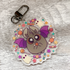 The CandyBat Keychain from Bindlewood Shop is a round acrylic charm featuring a cute cartoon bat with big eyes and purple wings holding candy. Inspired by Amanda Louise Spayd, the illustration shows the bat surrounded by colorful candies, stars, and tiny white ghosts on a light wood background.