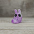 Plasma Storm Chenille, a small purple bunny-like resin figure by Bindlewood Shop and Amanda Louise Spayd, sits on a wooden surface with large black eyes and raised front paws. The background is blurred light brown wood.