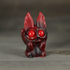 The Midnight Cranberry Nettlepup by Bindlewood Shop is a small red and black figurine with large round eyes and flame-like ears, sitting on a gray surface with glittery red swirl accents and an adorably surprised expression.
