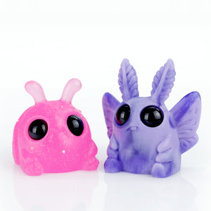 The Bindlewood Shop's Grape Jelly Swirl Edison and Neon Glitterberry Weebeetle Set features a purple figure with large eyes and wing-like details beside a pink one with antennae. Both have a smooth, glossy texture against a white background.