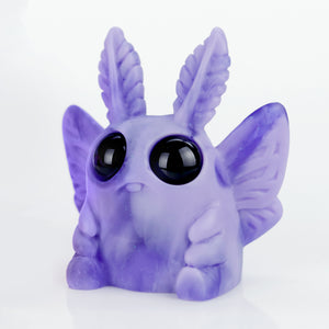 The Grape Jelly Swirl Edison by Amanda L Spayd is a small, cute pastel purple figure of a moth-like creature. It has large shiny black eyes, leaf-shaped antennae, stubby wings, and a chubby body resting on a smooth white surface.