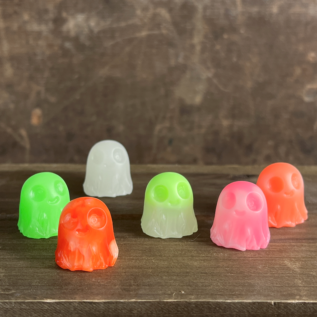 Six small, translucent Giddy Ghost Glow figurines from Bindlewood Shop—ghost-shaped collectibles in green, white, orange, and pink—are arranged on a wooden surface against a softly blurred brown background. Inspired by Chris Ryniak’s whimsical designs, these playful figures are sure to add a charming glow to any collection.