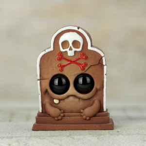 The "Gingerdead Hedston" by Chris Ryniak is a cute, handpainted resin figure resembling a cracked tombstone with large black eyes, a small smile, and skeletal arms. It displays a white skull with crossed red bones on its head against a neutral background.