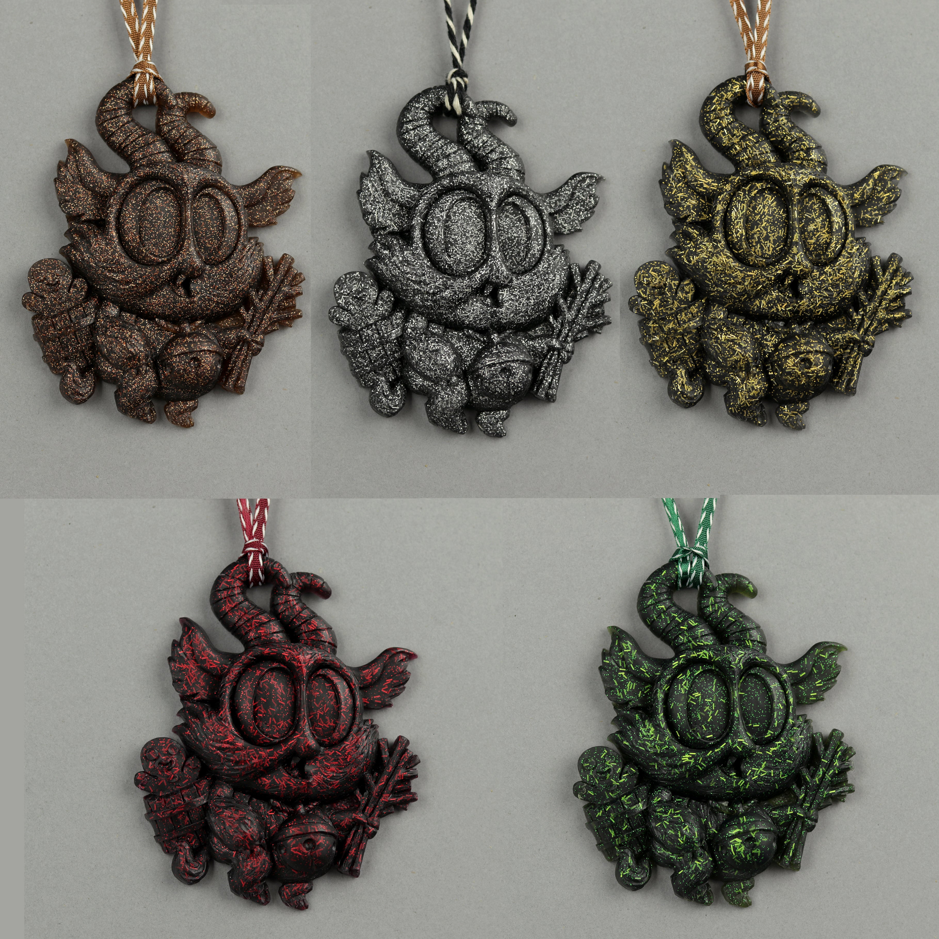 Five Glitter Krampus Ornaments by Bindlewood Shop—textured pendants shaped like horned creatures with big eyes, each in a different metallic hue—are displayed on a gray background with matching cords, sparkling like tiny resin treasures.