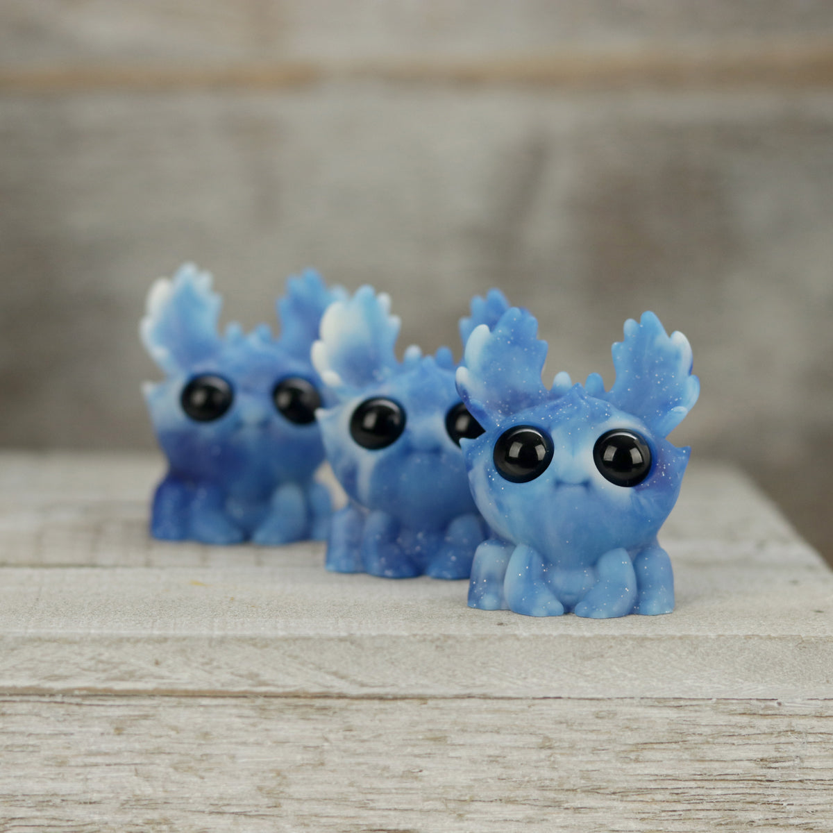 Three small blue Sunshower Moose resin figures by Bindlewood Shop sit on a wooden surface, featuring large black eyes and white-tipped antlers, set against a blurred wood background.
