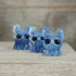 Three small blue Sunshower Moose resin figures by Bindlewood Shop sit on a wooden surface, featuring large black eyes and white-tipped antlers, set against a blurred wood background.