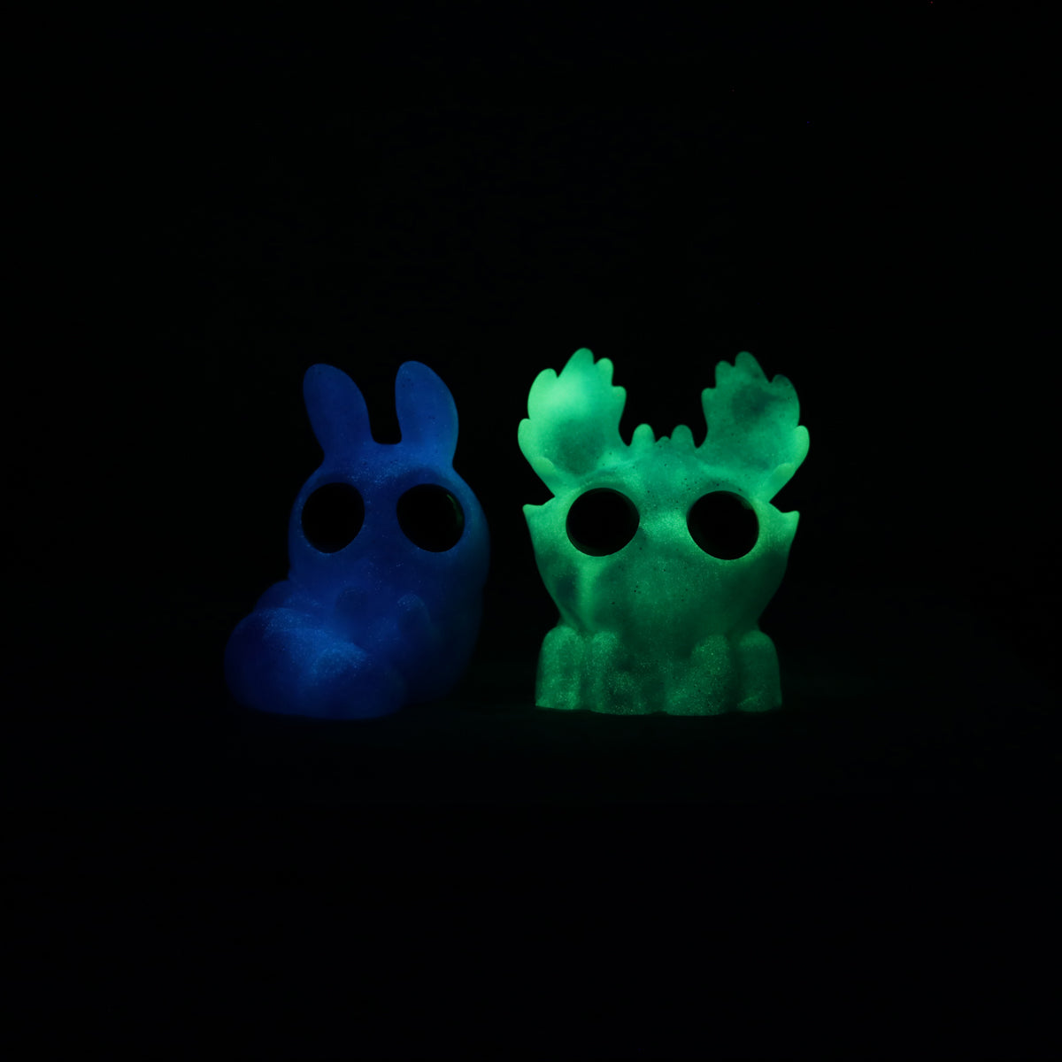 Two small, glowing resin figures from Bindlewood Shop are seen in the dark: Sunshower Moose with antler-like features and Plasma Storm Chenille with bunny-like ears, both from the Sunshower Moose & Plasma Storm Chenille Set.