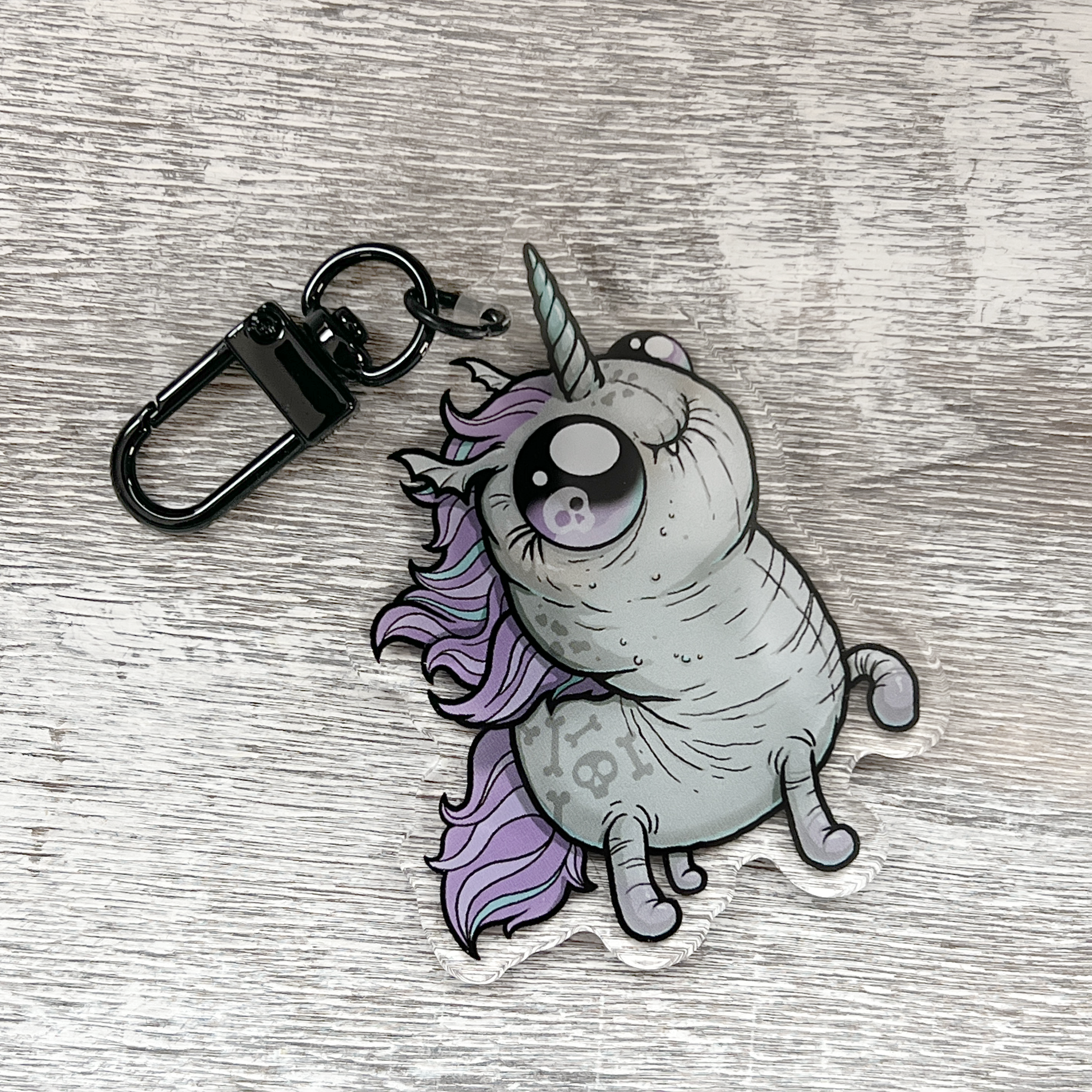 Presenting the Goth Ponycorn Keychain by Bindlewood Shop—a double-sided acrylic charm portraying a cartoon unicorn with a large eye, gray body, and purple mane and tail, lying on its back against a textured gray wooden background—perfect for fans of unique unicorn accessories.