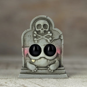 The Grayve Hedston by Chris Ryniak is a charming, handpainted resin figure featuring a cute tombstone with big black eyes, blushing cheeks, and a skull and crossbones design. It brings giggles to the graveyard against a blurred neutral background.