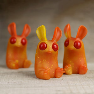 Three Sparkle Flame Peppercorn resin rabbit figurines with bright red eyes and yellow interior details, positioned on a wooden surface with a soft focus background. Brand Name: Bindlewood Shop