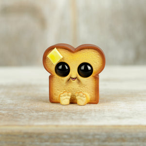 Handpainted Buttered Toastboy by Chris Ryniak is a cute cartoon bread slice with big shiny eyes, a small smile, tiny feet, and a butter pat on top, standing on light wood—a perfect hand-painted resin figure.