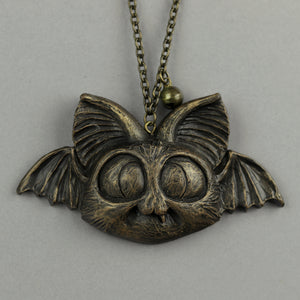 Painted Bat Ornaments by Bindlewood Shop feature a bronze pendant with a stylized bat face, large ears, wide eyes, and wing-like details—miniature bat sculptures inspired by Chris Ryniak, shown on a gray background.
