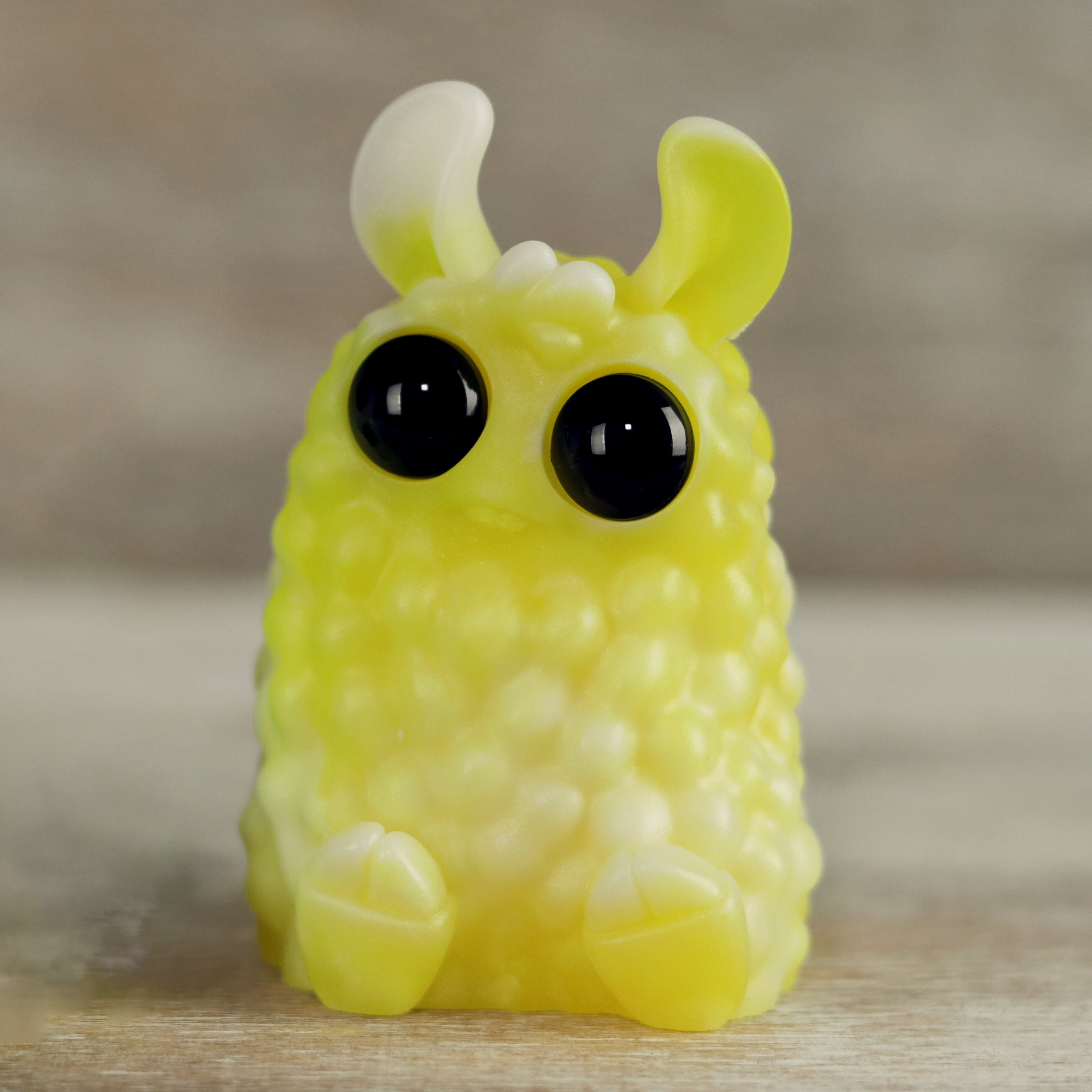The Lemon Meringue PomPom from Bindlewood Shop is a small yellow and white resin figure by Amanda Louise Spayd, featuring big black eyes, two rounded ears, and tiny feet, sitting on a light surface with a blurred background.