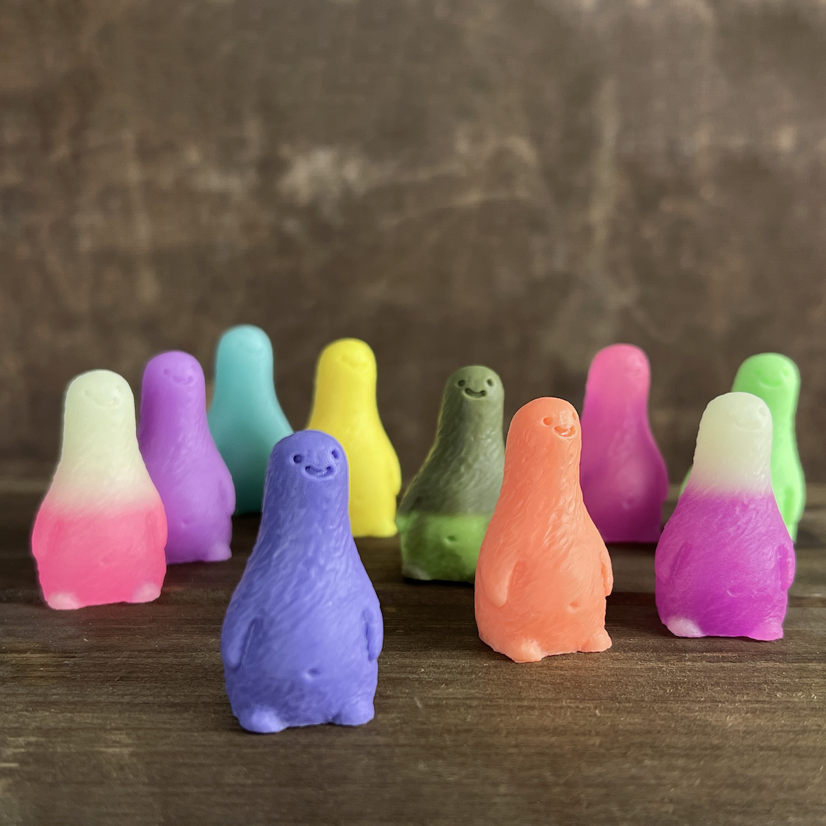 Two small, colorful, smiling Little Guy figurines from the Bindlewood Shop Little Guy 2-pack—collectible figures inspired by Chris Ryniak—stand on a wooden surface against a blurred brown background.