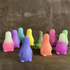 Two small, colorful, smiling Little Guy figurines from the Bindlewood Shop Little Guy 2-pack—collectible figures inspired by Chris Ryniak—stand on a wooden surface against a blurred brown background.