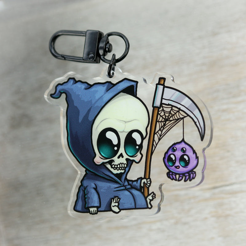 The Lil' Reaper Keychain by Bindlewood Shop features Chris Ryniak’s adorable charm in a blue hood, holding a scythe with a purple spider, both sporting big expressive eyes for added appeal.
