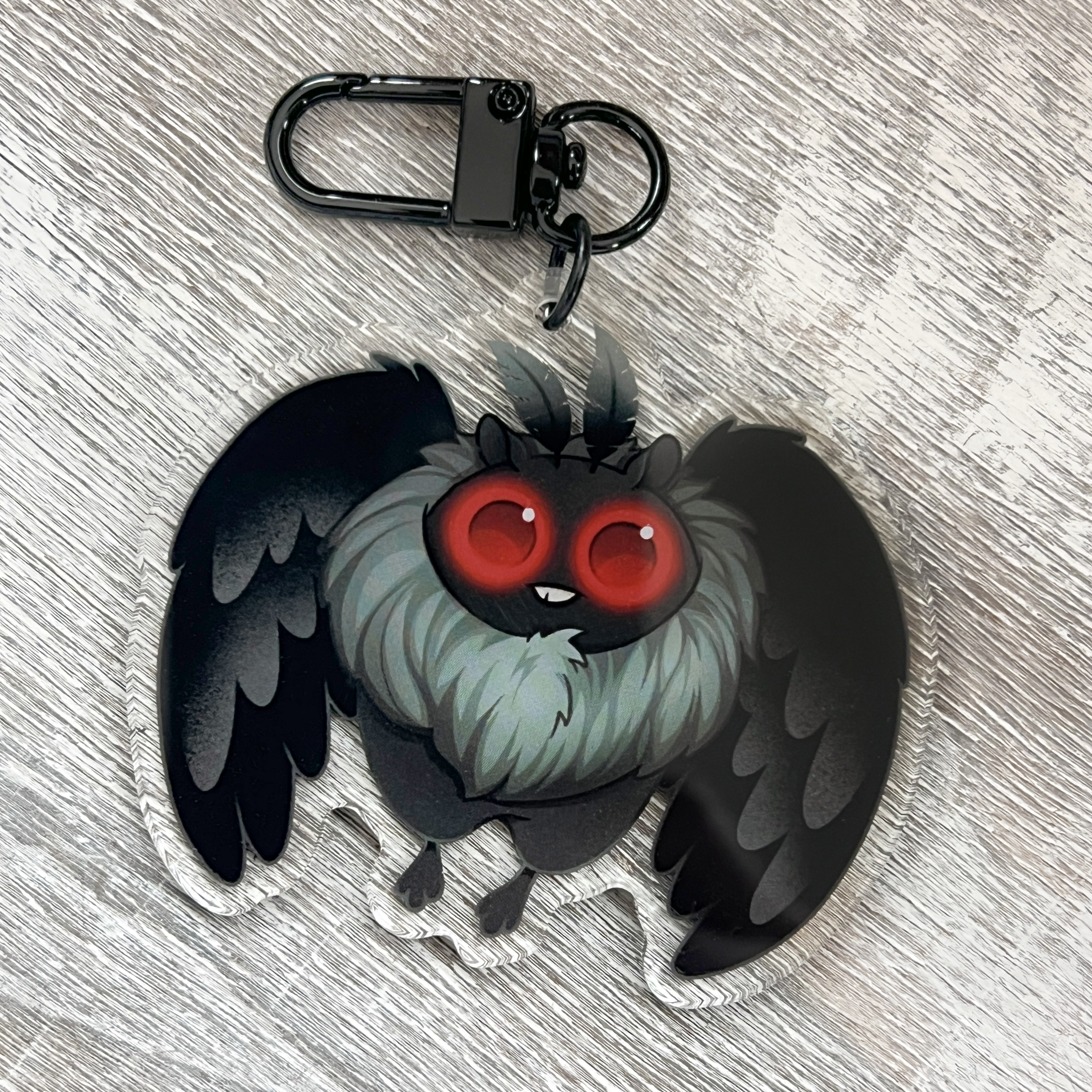 The Bindlewood Shop Mothman Keychain features a double-sided acrylic charm depicting a cartoon bat-like creature with big red eyes, fluffy grey fur, and black wings. Presented on a light wood surface, this keychain includes a coated metal clip for easy attachment.