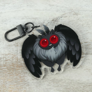 A whimsical keychain featuring a stylized "Mothman" Acrylic Charm with large red eyes and fluffy grey and black feathers from Bindlewood Shop on a rustic white wooden background.