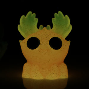 The Electric Peach Moose by Bindlewood Shop is a whimsical resin figure that glows in the dark. It features large eye holes and leaf-like green ears on its orange head, evoking a playful moose. The speckled texture adds charm, while the darkness highlights its glow-in-the-dark effect.