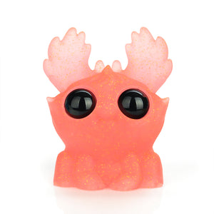 The Electric Peach Moose by Bindlewood Shop is a charming toy with antler-like ears, large black eyes, and a glitter-speckled pink surface that shimmers. Its glow-in-the-dark feature enhances its whimsical appeal.