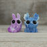 The Bindlewood Shop Sunshower Moose & Plasma Storm Chenille Set features two small, glittery resin figures with large black eyes—a purple bunny and a Caribbean Blue Dashi-inspired moose—side by side on a wooden surface.
