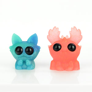 Two cute resin figures are shown on a white background from the Bindlewood Shop. On the left is a Caribbean Blue Dashi with pointy ears, and on the right is an Electric Peach Moose with rounded antler-like shapes. Both have large glossy black eyes and a whimsical appearance.