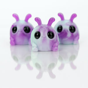 Three small Plasma Storm Weebeetle figures by Chris Ryniak, with big black eyes and two antennae, stand on a reflective white surface, featuring purple and white colors in a whimsical cartoonish style.