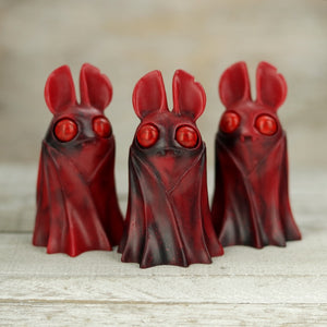 Three red, small resin figures in cloaks with large eyes and pointed ears stand together. Crafted by Amanda Louise Spayd, they are named "Cranberry Jasper Belfry" by Bindlewood Shop, set against a wooden textured backdrop enhancing their charm.